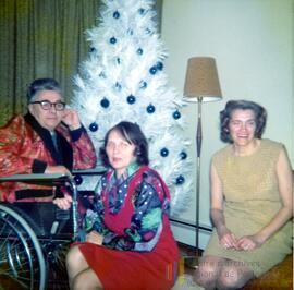 Un homme en fauteuil roulant et deux femmes devant un sapin de Noël blanc