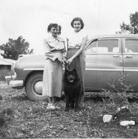 Deux femmes avec un chien noir en laisse près d’une voiture