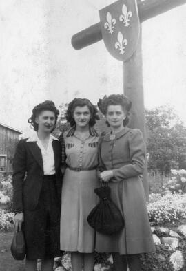 Trois femmes dehors devant une grande croix décorée de trois fleurs de lys