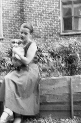 Femme assise sur un clôture devant un bâtiment tenant un chien dans ses bras