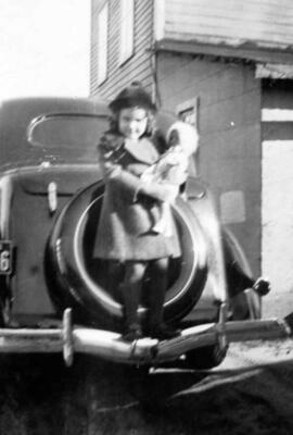Une enfant (peut-être Louise) avec une poupée derrière une voiture