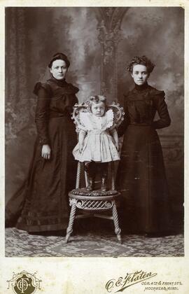 Photographie de deux femmes debout et une fillette debout sur une chaise