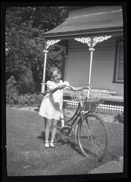 Une petite fille non identifiée à côté d’une bicyclette devant une maison
